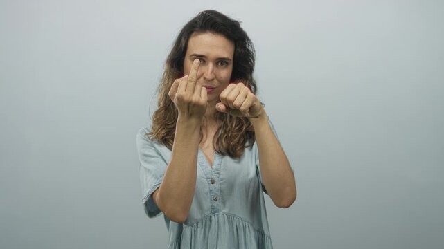 Woman wearing a denim dress raises a middle finger with direct gaze and clenches a fist in a grey studio; defiance.