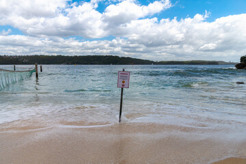 Warning sign on the sand at Shark Beach in Nielsen Park, Sydney Harbour, Australia, alerting visitors to hazards in the water near the shoreline.