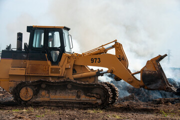 A man in a yellow track loader drives by a debris pile on fire with smoke rising in the air at a demolition site during the summer.
