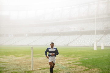 Rugby player is running on marked grass pitch inside large stadium, holding ball toward camera