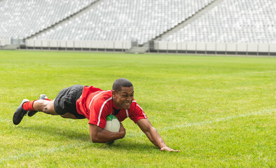 Rugby player is diving and clutching white-and-green ball on marked grass pitch near stands