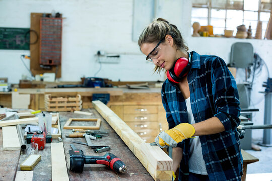 Female woodworker wearing safety goggles and gloves planing long timber at shop bench, copy space