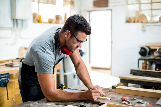 Male carpenter leaning over bench in workshop, measuring wood with tape, wearing safety glasses