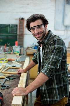 Male woodworker positioning long plank on cluttered workbench at workshop, wearing safety glasses