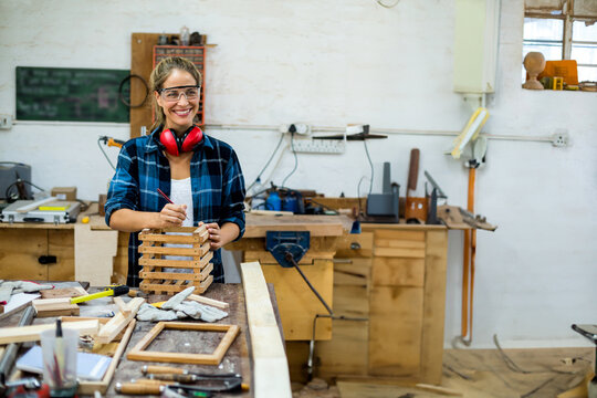 Adult woman standing and marking stacked slats at bench, holding pencil, wearing safety glasses
