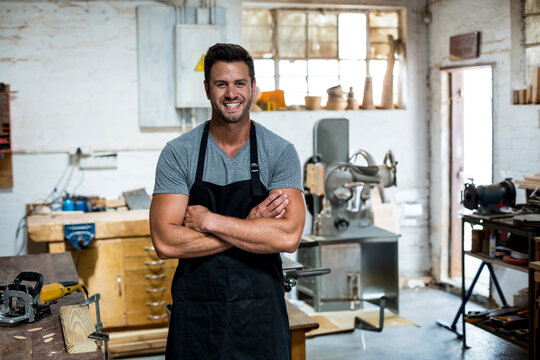 Male carpenter standing arms crossed in workshop by bench with vise and power tools wearing apron