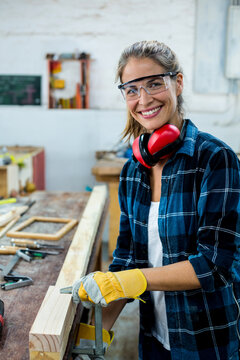 Female carpenter working at bench in shop wearing goggles red earmuffs yellow gloves clamping beam