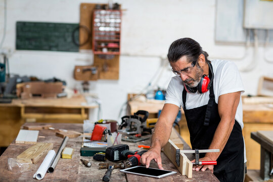 Mature carpenter in black apron leaning over clamped board at bench using tablet, drill, copy space