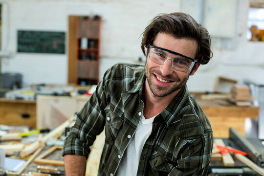 Male woodworker leaning on cluttered workbench in busy workshop with wood planks, clamps, smiling