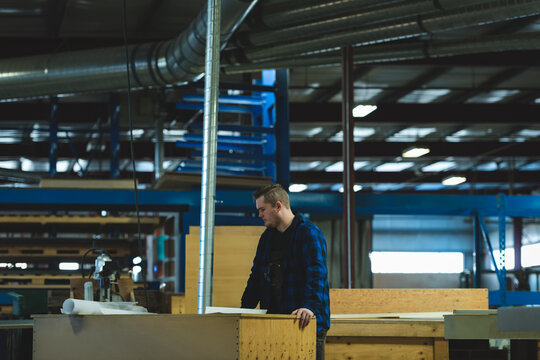 Large wooden workbench is holding stacked plywood and rolled plans in warehouse, blue racks