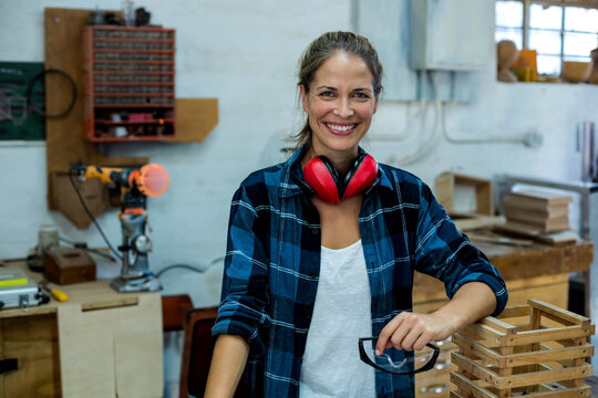 Female woodworker smiling at camera in workshop wearing plaid shirt with red earmuffs holding specs
