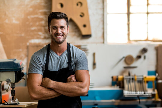 Male woodworker wearing black apron, standing arms crossed in workshop, drill press nearby