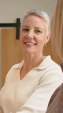 Vertical video: Smiling woman answering offscreen friend in kitchen, cream top and stud earring
