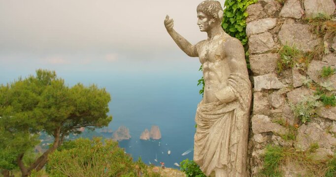 Ancient stone statue of Emperor Augustus stands against a rocky wall overlooking the distant Faraglioni sea stacks of Capri Italy