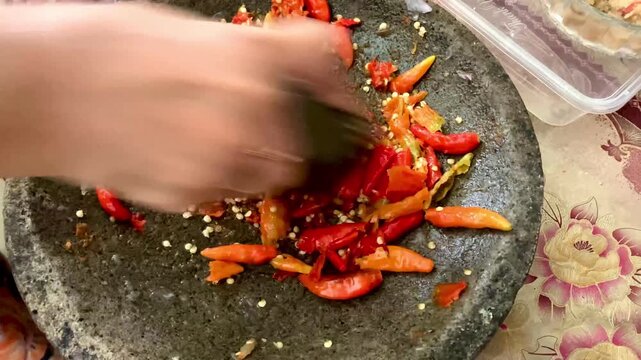hand grinding red chili peppers with stone mortar and pestle to make traditional spicy sambal sauce