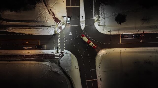 Top-down aerial of a well-lit road intersection in Vilnius at night, with snow-covered sidewalks, marked lanes, and a red bus turning through the junction.