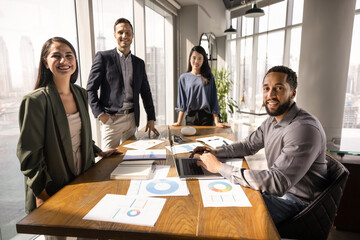 Diverse business team of happy young coworkers meeting at conference table with paper reports for analysis work, looking at camera, smiling. Marketing agency staff posing for group portrait