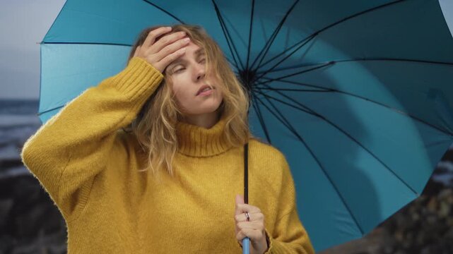 Woman holds umbrella looking anxious on a beach while her blonde hair frames a young face clad in a turtleneck mustard sweater beneath a blue canopy during rain.
