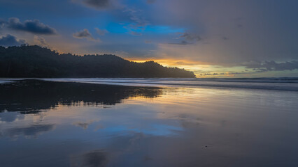 Fototapeta premium Evening after sunset. A tropical island. The sky is highlighted in gold near the horizon. Purple clouds. Ocean waves are spreading across the sandy beach. Reflection on wet, smooth sand. Long exposure