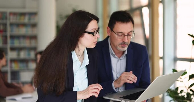 Middle-aged male employee holding laptop, explains online task, reviewing project information, clarify new software to female colleague during meeting in office. Discussion, mentorship, teamwork,