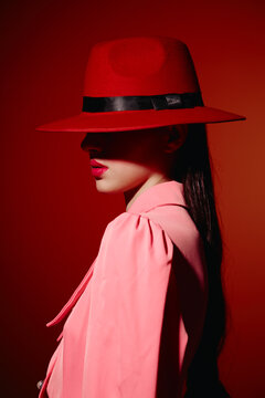 Stylish woman wearing a wide-brimmed red hat and pink coat with bright red lipstick poses against a red background. Dramatic shadow covers her face for a creative beauty concept.