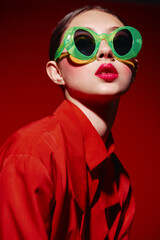 Fashion creative woman with vibrant red lipstick wears oversized green sunglasses and a red jacket...