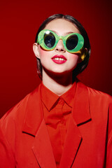 Naklejka premium Stylish woman with bright red lipstick wearing oversized green sunglasses and a red outfit against a vibrant red background. Concept of beauty, fashion, creative style, and modern trends.