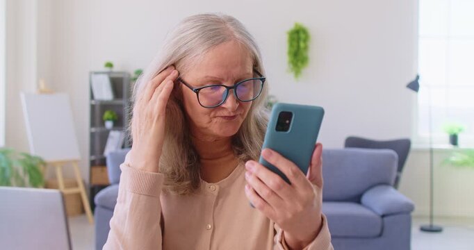 Older woman in glasses using smartphone at home, looks at device screen with concerned expression, experiencing problems with modern gadget, lack of understanding, need help with mobile application