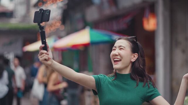 Happy young Asian woman vlogger recording a travel video with a smartphone on a selfie stick while exploring a city street. Modern influencer and digital lifestyle concept.