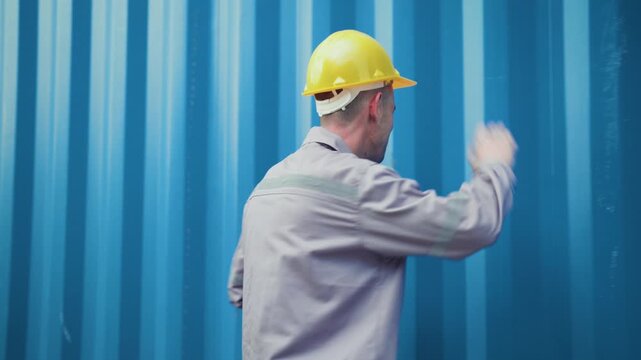 Frustrated industrial worker in a safety helmet shouting and reacting angrily while looking at his smartphone in front of a blue shipping container. Concept of job loss, layoff, and workplace crisis.