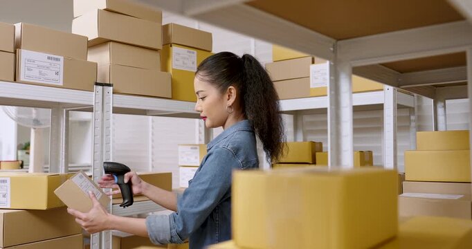 Asian woman small business owner holding parcel box while scanning shipping label, reviewing inventory location, managing e-commerce logistics workflow, online store shipment inside warehouse storage