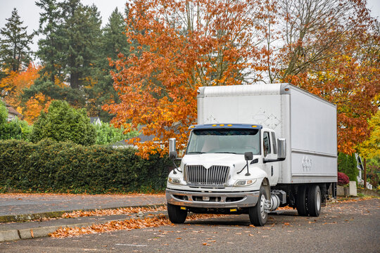 Industrial grade day cab white middle duty rig semi truck with box trailer standing on the local city street with autumn trees on the background