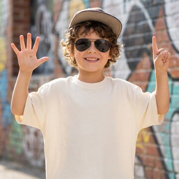 Cool boy model in sunglasses, baseball cap, wearing oversized natural t-shirt, pants, holding up six fingers, stands by graffiti wall. Youth crewneck short sleeve t-shirt mockup, summer city street