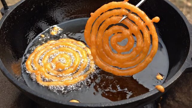 Close up of golden jalebi being deep fried in boiling oil as a cook uses a metal tool to lift and turn the spiral shaped indian dessert
