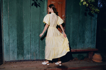 Woman in a bright yellow vintage dress walks along a weathered green wall, creating a nostalgic outdoor fashion moment with soft lighting and graceful movement.