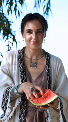 A beautiful brunette in a bohemian style relaxes outdoors in the summer, holding a large slice of watermelon against a green forest backdrop.