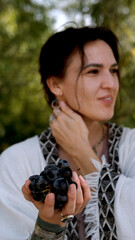 A beautiful brunette in a bohemian style relaxes outdoors in summer against a backdrop of a green forest, holding a large bunch of juicy black grapes