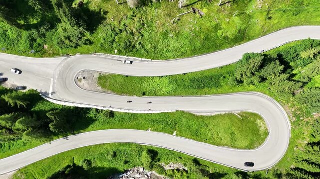 Top down drone view of S road, S curve in the Italian dolomites, some cars and motercycles are driving on the road, taking curves. Road surrounded by greenery, grass and trees, beautiful nature. 