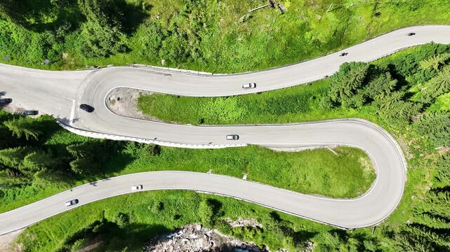 Top down drone view of S road, S curve in the Italian dolomites, some cars and motercycles are driving on the road, taking curves. Road surrounded by greenery, grass and trees, beautiful nature. 
