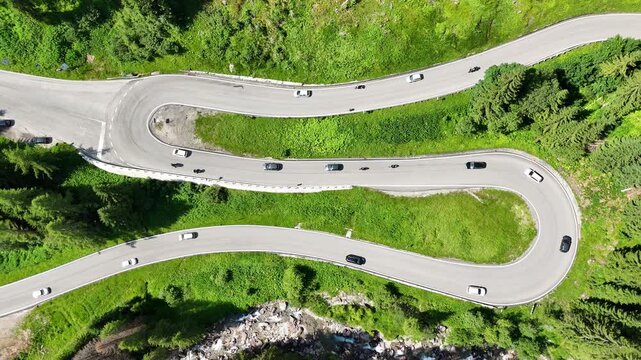Top down drone view of S road, S curve in the Italian dolomites, some cars and motercycles are driving on the road, taking curves. Road surrounded by greenery, grass and trees, beautiful nature. 
