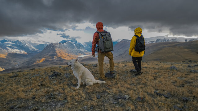 A couple in bright rain jackets and a white dog stand on a mountain ridge during an autumn hiking trip, looking at a dramatic landscape of distant snow-capped peaks under heavy, cloudy skies.