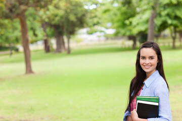 Fototapeta premium Female student standing on right of park wearing blue shirt, holding textbooks, smiling, copy space