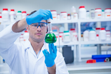 Glass flask with bright green solution is being pipetted on lab bench, shelves visible