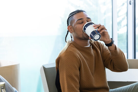 African American man in brown quilted top sipping blue-accented cup in lounge by big windows