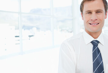 Fototapeta premium Mid-adult man standing in office by glass windows wearing white shirt navy-blue tie, copy space