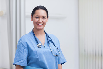 Female nurse in blue scrubs standing, smiling, facing camera in clinic with stethoscope and folder © wavebreak3