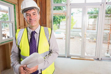 Senior man wearing white hard hat and hi-vis vest holding rolled blueprints inside renovated room