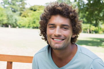 Adult man smiling while sitting on wooden park bench in sunny park wearing light blue T-shirt