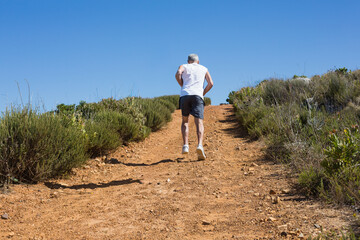 Runner is running uphill on dry rocky trail with low green shrubs under blue sky