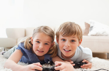 Brother and sister children lying on thick light-colored rug at home, holding wired controllers